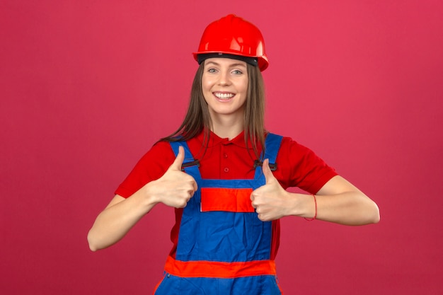 Young woman in construction uniform and red safety helmet happy looking showing thumbs up standing on dark pink background