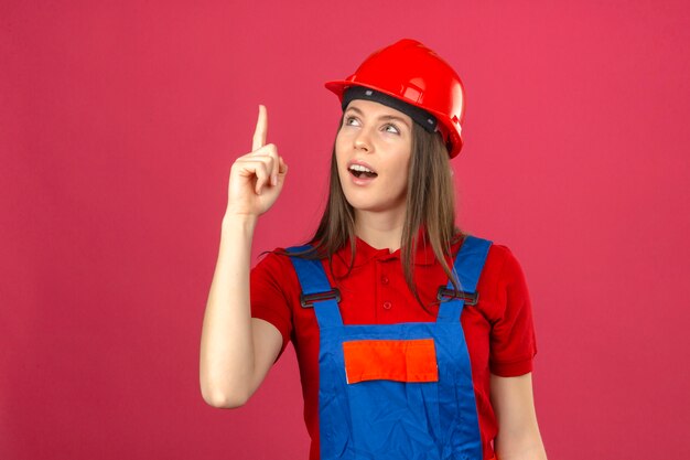 Young woman in construction uniform and red safety helmet having great idea pointing finger up on dark pink background