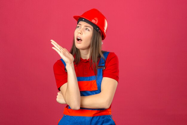 Young woman in construction uniform and red safety helmet looking sideway thinking an idea on dark pink background