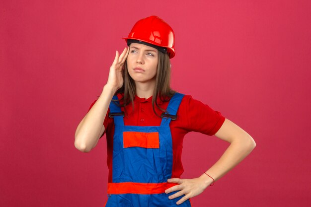 Young woman in construction uniform and red safety helmet looking sideways touching her head having a headache standing on dark pink background