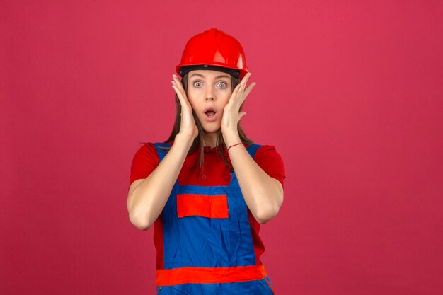 暗いピンクの背景にカメラを見て彼女の顔に触れるショックを受けた建設の制服と赤い安全ヘルメットの若い女性