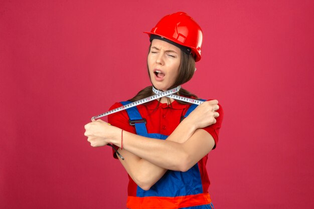 Young woman in construction uniform and red safety helmet shouting and choking herself with measurement tape on dark pink background