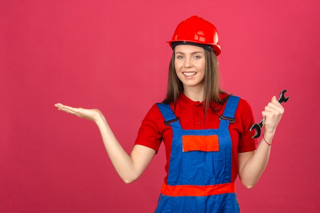 Young woman in construction uniform and red safety helmet smiling cheerful presenting and pointing with palm of hand and holding wrench on dark pink background