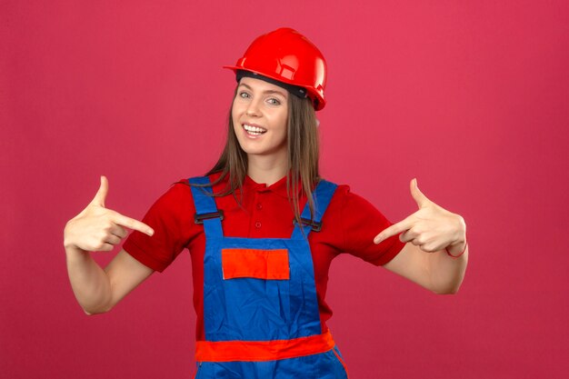 Young woman in construction uniform and red safety helmet smiling and pointing at herself looks joyful standing on dark pink background