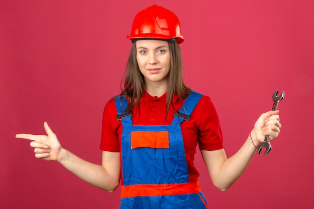 Free photo young woman in construction uniform and red safety helmet smiling and pointing with finger to the side and holding adjustable wrench on dark pink background