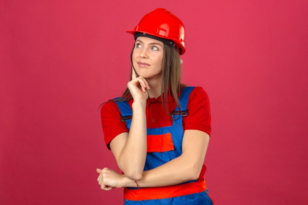 Молодая женщина в строительной форме и красный защитный шлем, улыбаясь, думая, новая идея, стоя на темно-розовом фоне