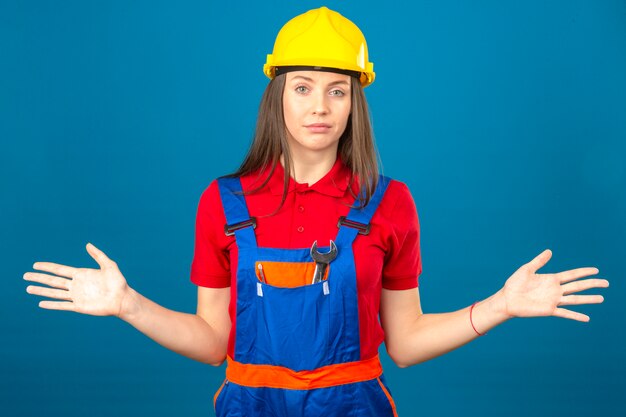 Young woman in construction uniform and yellow safety helmet clueless and confused expression with arms and hands raised standing on blue background