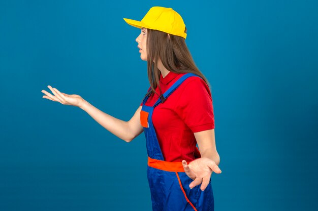 파란색 배경에 서 shrugging와 몸짓 건설 유니폼과 노란색 안전 헬멧에 젊은 여자
