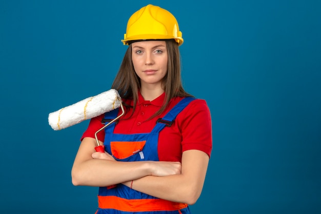 青の背景に組んだ腕でペイントローラーの地位を保持している建設の制服と黄色の安全ヘルメットの若い女性