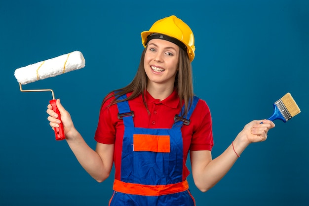 Young woman in construction uniform and yellow safety helmet smiling  holding paint roller and brush in hands standing on blue isolated background
