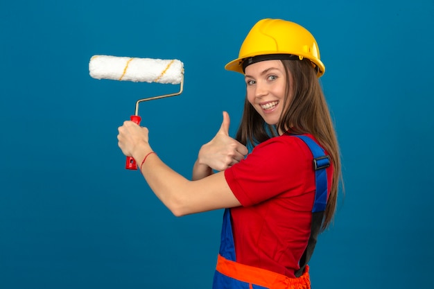 Young woman in construction uniform and yellow safety helmet smiling showing thumb up and holding paint roller in hand on blue isolated background