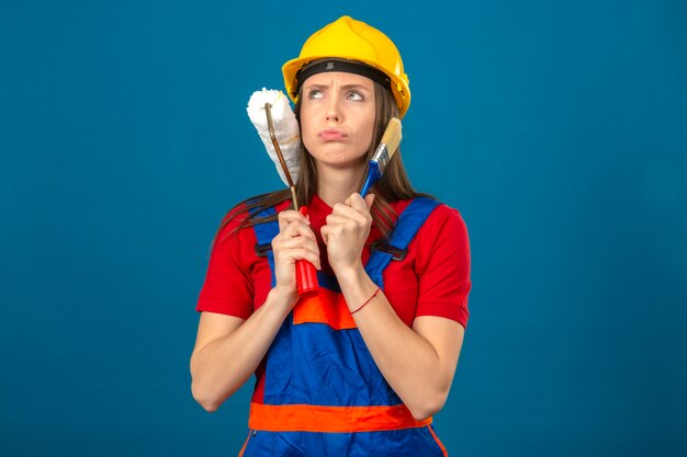 Young woman in construction uniform and yellow safety helmet thinking with serious face pensive expression holding paint roller and brush standing on blue background