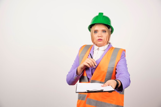 Young woman contractor with green helmet and clipboard on white.