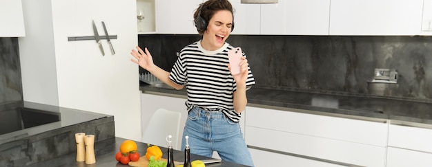Foto gratuita giovane donna che cucina il pasto in cucina ascoltando musica con le cuffie wireless che canta