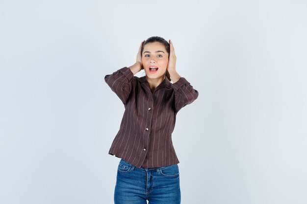 Young woman covering ears with hands, keeping mouth open in striped shirt, jeans and looking harried. front view.