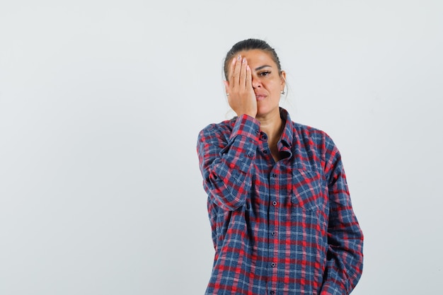 Young woman covering eye with hand in checked shirt and looking serious , front view.