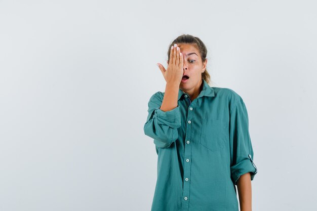 Young woman covering eye with hand in green blouse and looking surprised