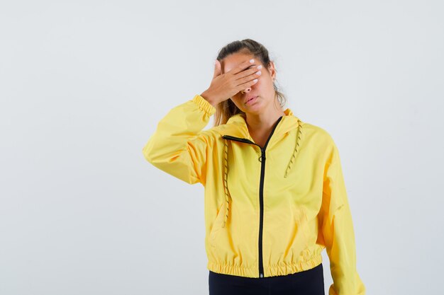 Young woman covering eyes with hand in yellow raincoat and looking bored