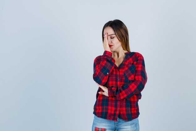 Young woman covering face with hand in checked shirt, jeans and looking annoyed. front view.