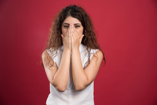 Young woman covering face with hands on red studio wall. 