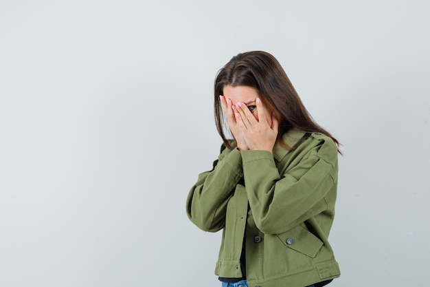 Free photo young woman covering face with hands while looking through fingers in green jacket , front view. space for text