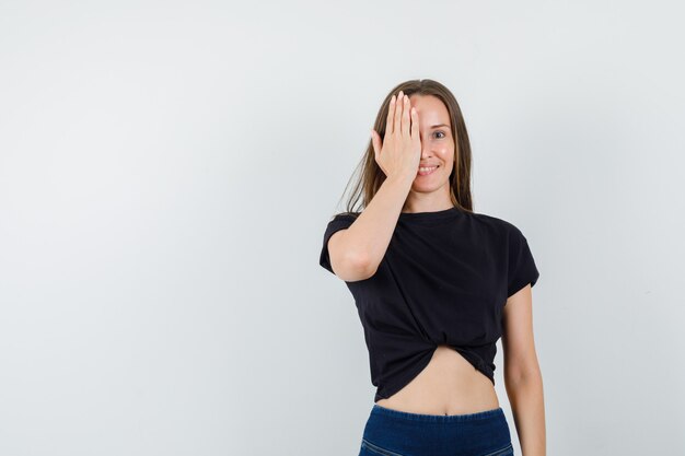 Young woman covering her hand on one eye in black blouse and looking joyful.