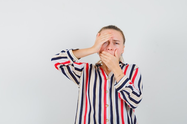 Young woman covering her mouth and head with hands in striped blouse and looking annoyed