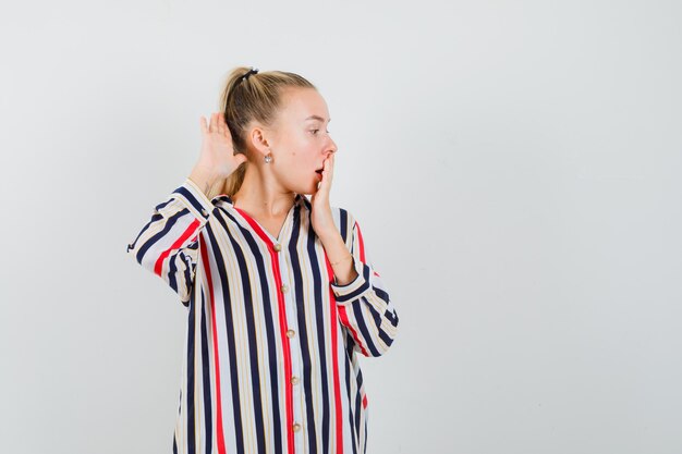 Young woman covering her mouth with hand and trying to hear something in striped blouse and looking surprised