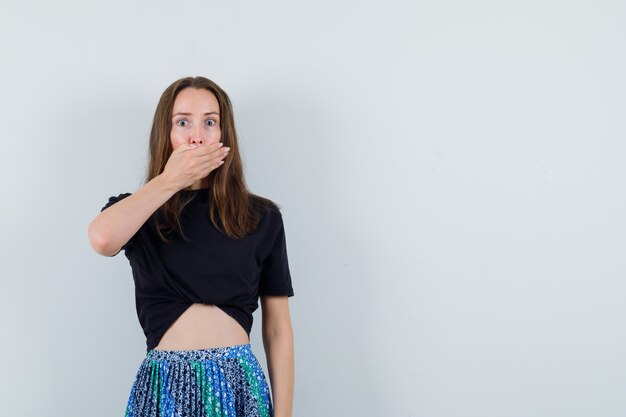 Young woman covering mouth with hand in black t-shirt and blue skirt and looking surprised