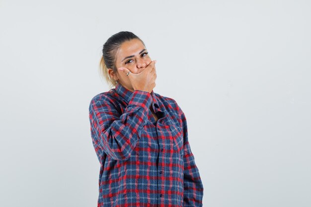 Young woman covering mouth with hand in checked shirt and looking pretty. front view.