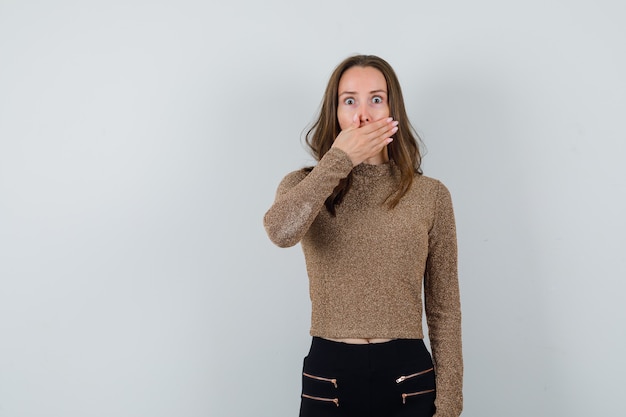 Young woman covering mouth with hand in gold gilded sweater and black pants and looking surprised