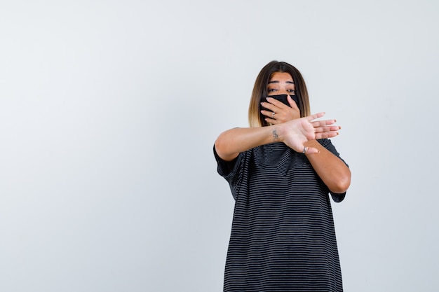 Young woman covering mouth with hand, showing stop sign in black dress, black mask and looking scared , front view.