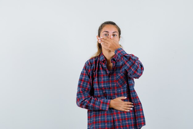 Young woman covering mouth with hand while holding hand on belly in checked shirt and looking tired