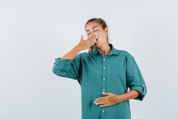 Young woman covering mouth with hand and yawning in green blouse and looking sleepy