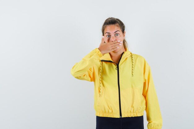 Young woman covering mouth with hand in yellow bomber jacket and black pants and looking surprised