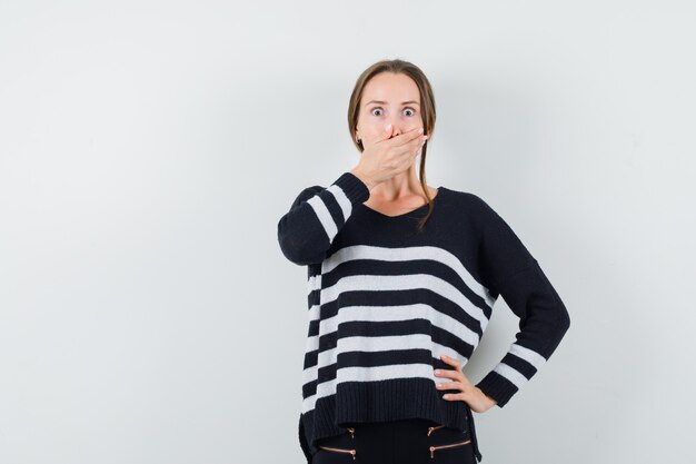 Young woman covering mouth with hands while holding hands on waist in striped knitwear and black pants and looking surprised