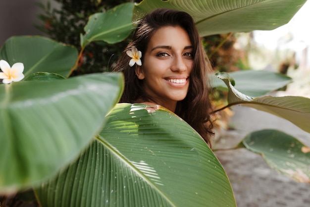 Young woman covers herself with huge green leaf and looks into camera with smile