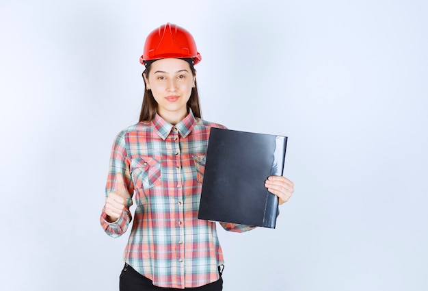 Free photo a young woman in crash helmet holding black folder and showing thumb up.