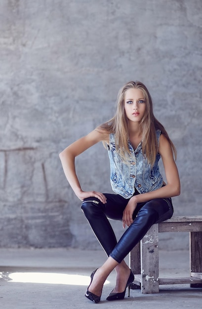 A young woman in denim jacket and black pants posing near concrete wall.