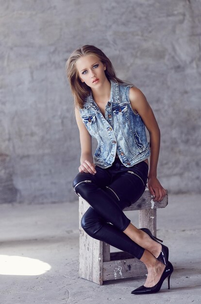 A young woman in denim jacket and black pants posing near concrete wall.