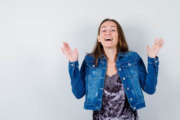 Young woman in denim jacket, dress raising hands, opening mouth and looking delighted , front view.