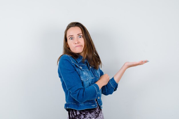 Young woman in denim jacket spreading palm aside and looking puzzled , front view.