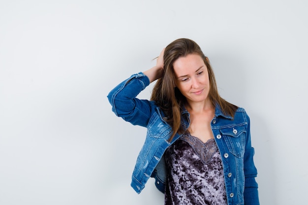 Young woman in denim jacket with hand on head, looking down and looking dreamy , front view.