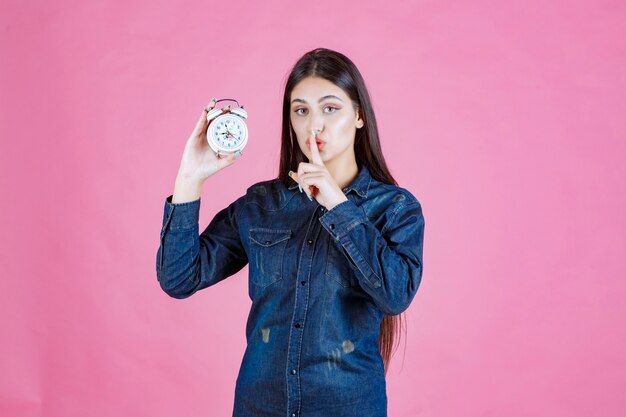 Young woman in denim shirt holding the alarm clock and making silence sign
