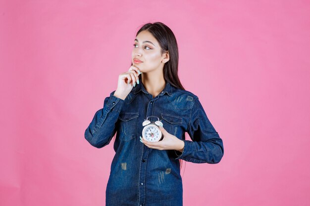 Young woman in denim shirt holding the alarm clock and thinking