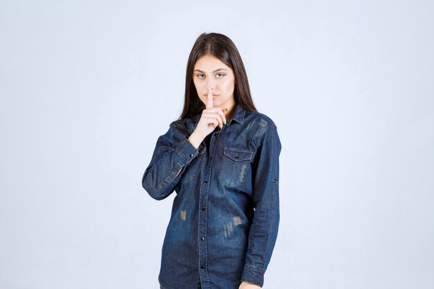 Young woman in denim shirt pointing at her mouth and asking to be quiet