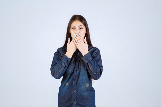 Young woman in denim shirt pointing at her mouth and asking to be quiet