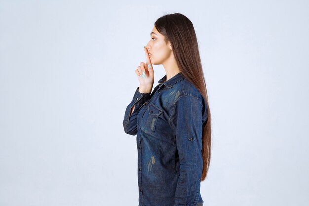 Young woman in denim shirt pointing at her mouth and asking to be quiet