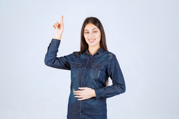 Young woman in denim shirt raising her hands up and pointing at something above