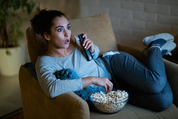 Young woman in disbelief watching TV while eating popcorn in the evening at home